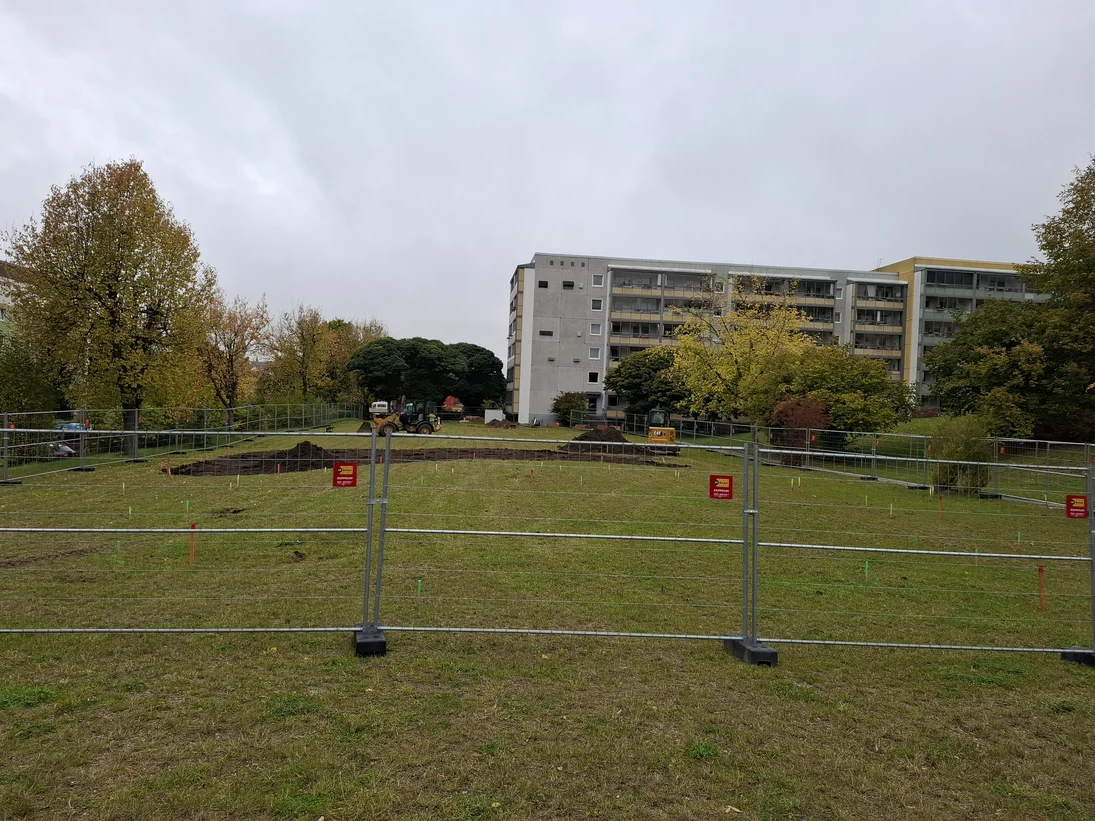 Zu sehen ist eine grosse Wiese auf der ein Bagger arbeitet. Im Vordergrund ist ein Bauzaun und im Hintergrund ein Wohnblock.