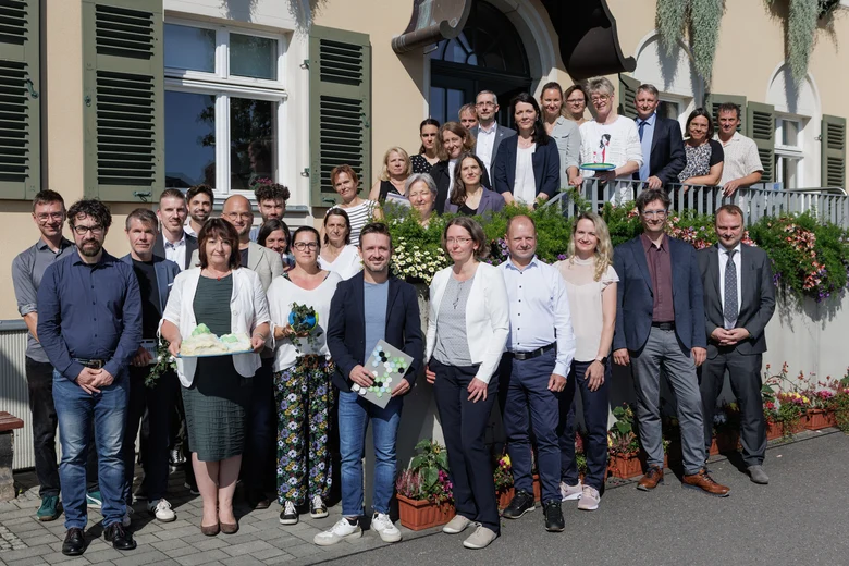 Gruppe aus Menschen die sich auf einer Treppe aufreihen, in die Sonne lächeln und Geschenke präsentieren