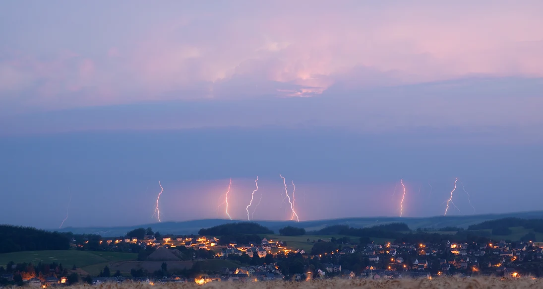 Fernblick auf eine Gemeinde, im Hintergrund dunkle Wolken und viele Blitze, die in die Erde einschlagen