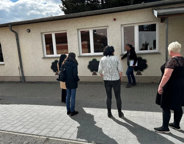 Planer und Verantwortliche besichtigen die Schule vor Ort: Personen schauen von außen auf das Schulgebäude, einen Flachbau; Personen mit dem Rücken zur Kamera; Mann zeigt etwas an der Wand