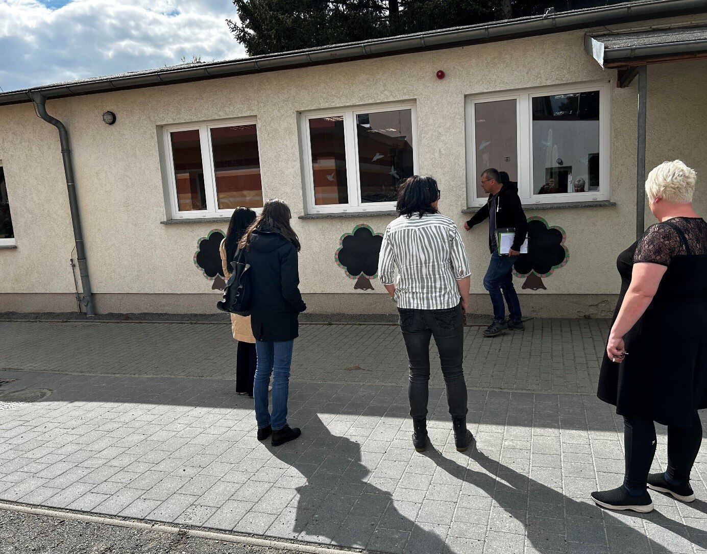 Planer und Verantwortliche besichtigen die Schule vor Ort: Personen schauen von außen auf das Schulgebäude, einen Flachbau; Personen mit dem Rücken zur Kamera; Mann zeigt etwas an der Wand