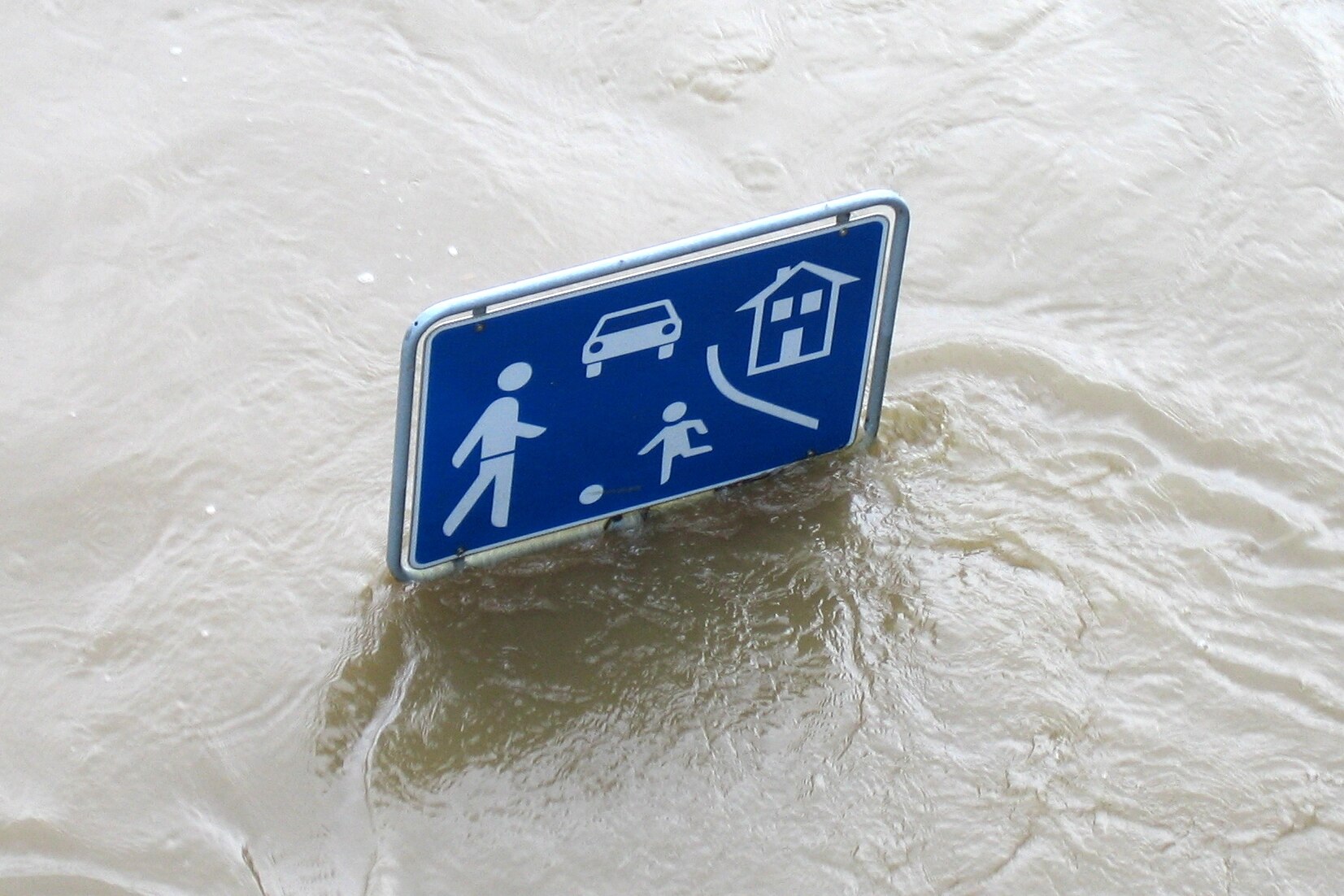 Foto von oben aufs Hochwasser und nur das Verkehrsschild "Verkehrsberuhigter Bereich" schaut noch raus (blaues rechteckiges Schild mit weißen ballspielenden Figuren und einem Auto).