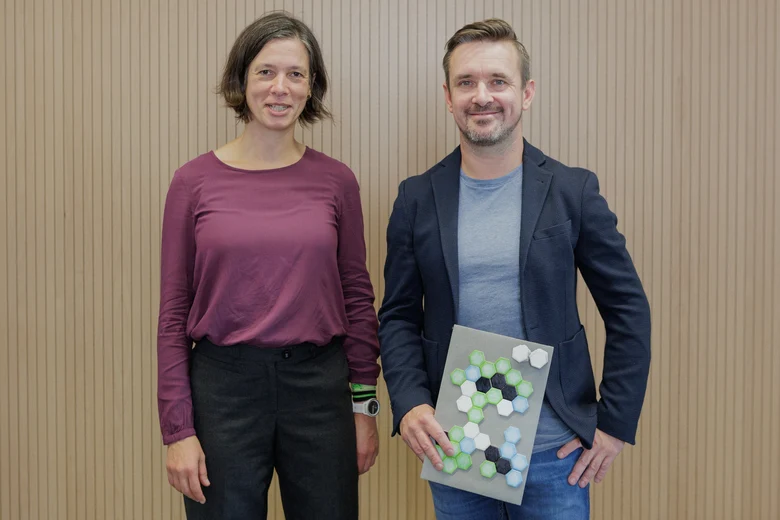 Eine Frau steht links und ein Mann rechts. Der Mann hält einen Preis in den Händen.