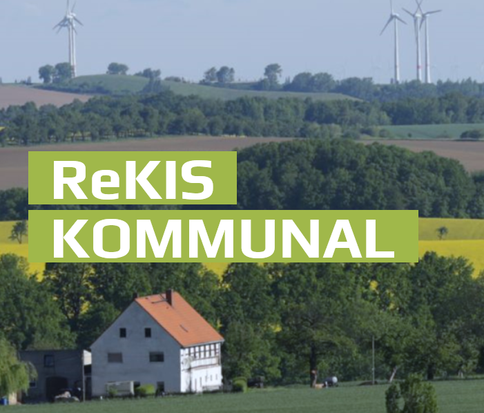 Schriftzug "ReKIS Kommunal" weiß auf hellgrünem Grund, links, dahinter ein Foto einer ländlichen Landschaft mit grünen Feldern und Bäumen, gelben Feld, Bauernhaus links und ganz hinten vor blauem Himmel Windräder