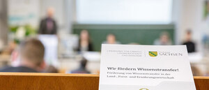 Symbolbild: Im Vordergrund ein Blattpapier mit einer Veranstaltungseinladung des LfULG; Hintergrund verschwommen, ein Verasntaltungsraum mit Zuhörern und Referent am Rednerpult lässt sich erahnen