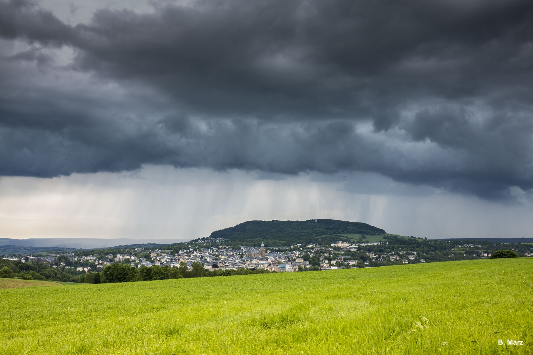 Helle Feld im Vordergrund, sächsische Kommune im Hintergrund darüber eine dunkle Regenwolke